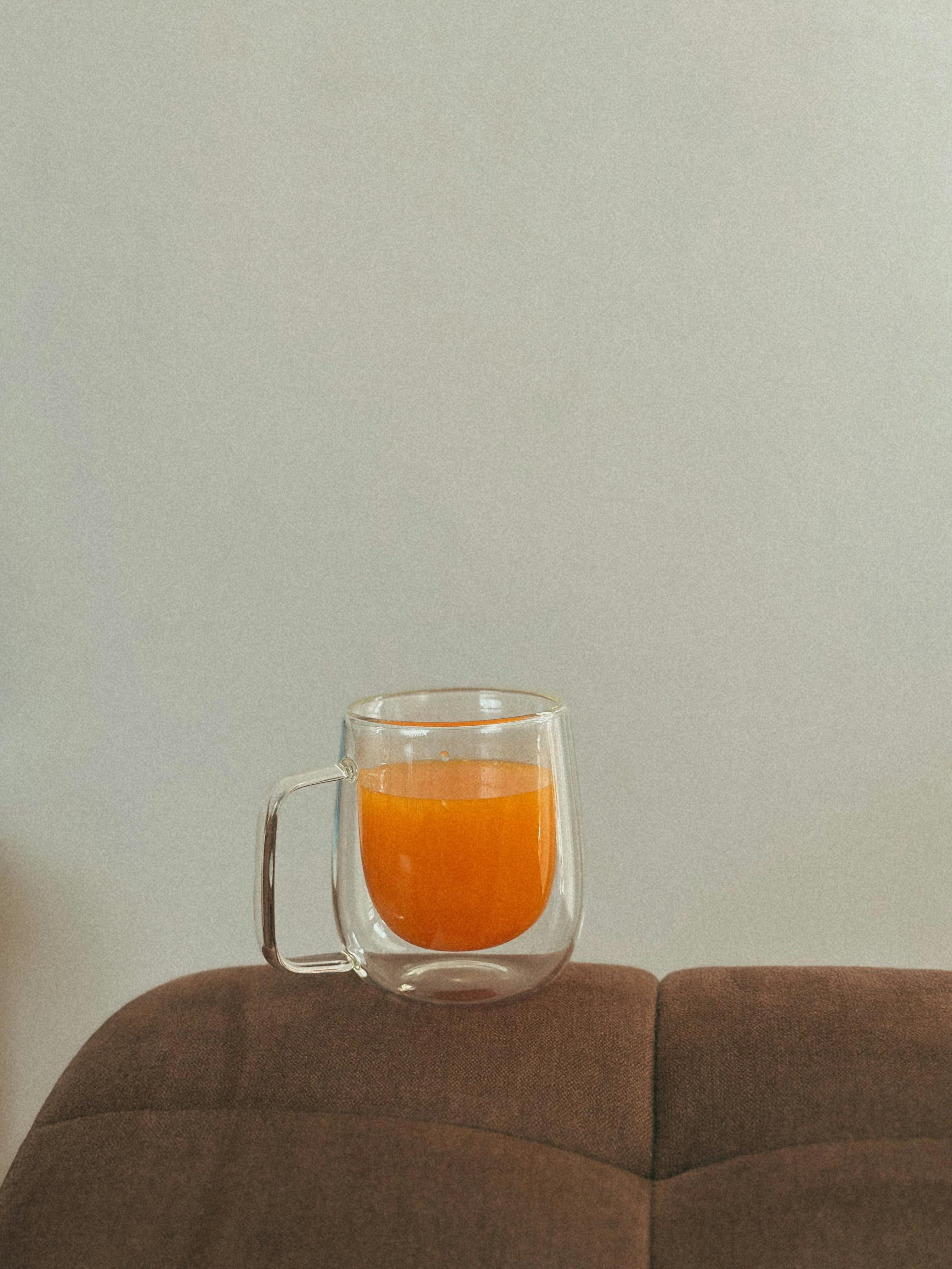 grátis Uma caneca de vidro transparente cheia de suco de laranja vibrante sobre uma almofada marrom, em ambiente interno. Foto profissional