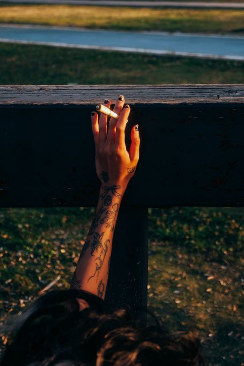Free A tattooed hand holds a cigarette near a wooden railing outdoors in golden sunlight. Stock Photo