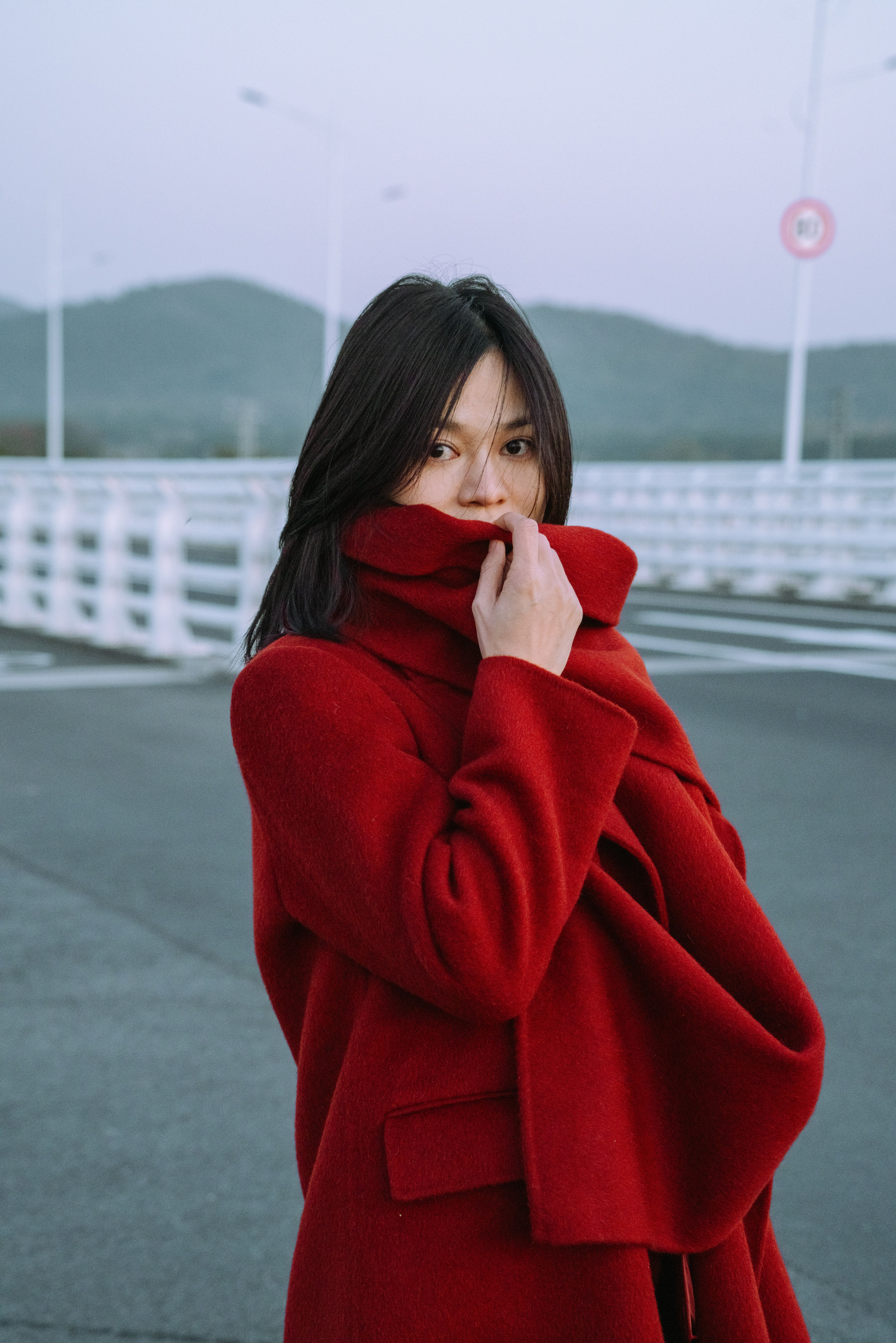 A woman in a vibrant red coat stands on a bridge outdoors with mountains visible in the background.