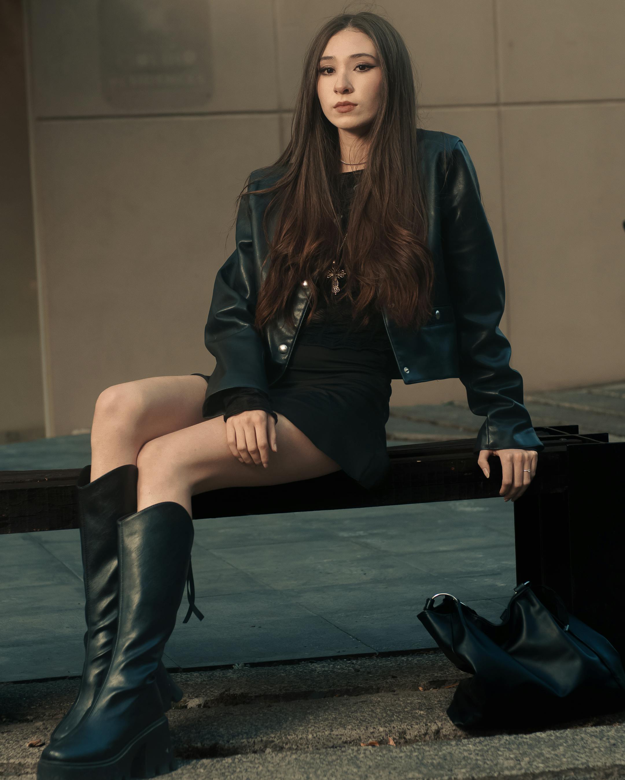 Free Confident woman wearing black leather jacket and boots sitting outdoors on a bench in an urban setting. Stock Photo