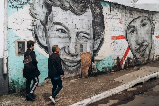Street art mural with iconic portraits on a wall in São Paulo, Brazil. Two people walk by.