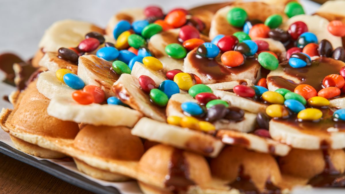 Close-up of a Belgian waffle topped with banana slices, colorful candies, and chocolate sauce.