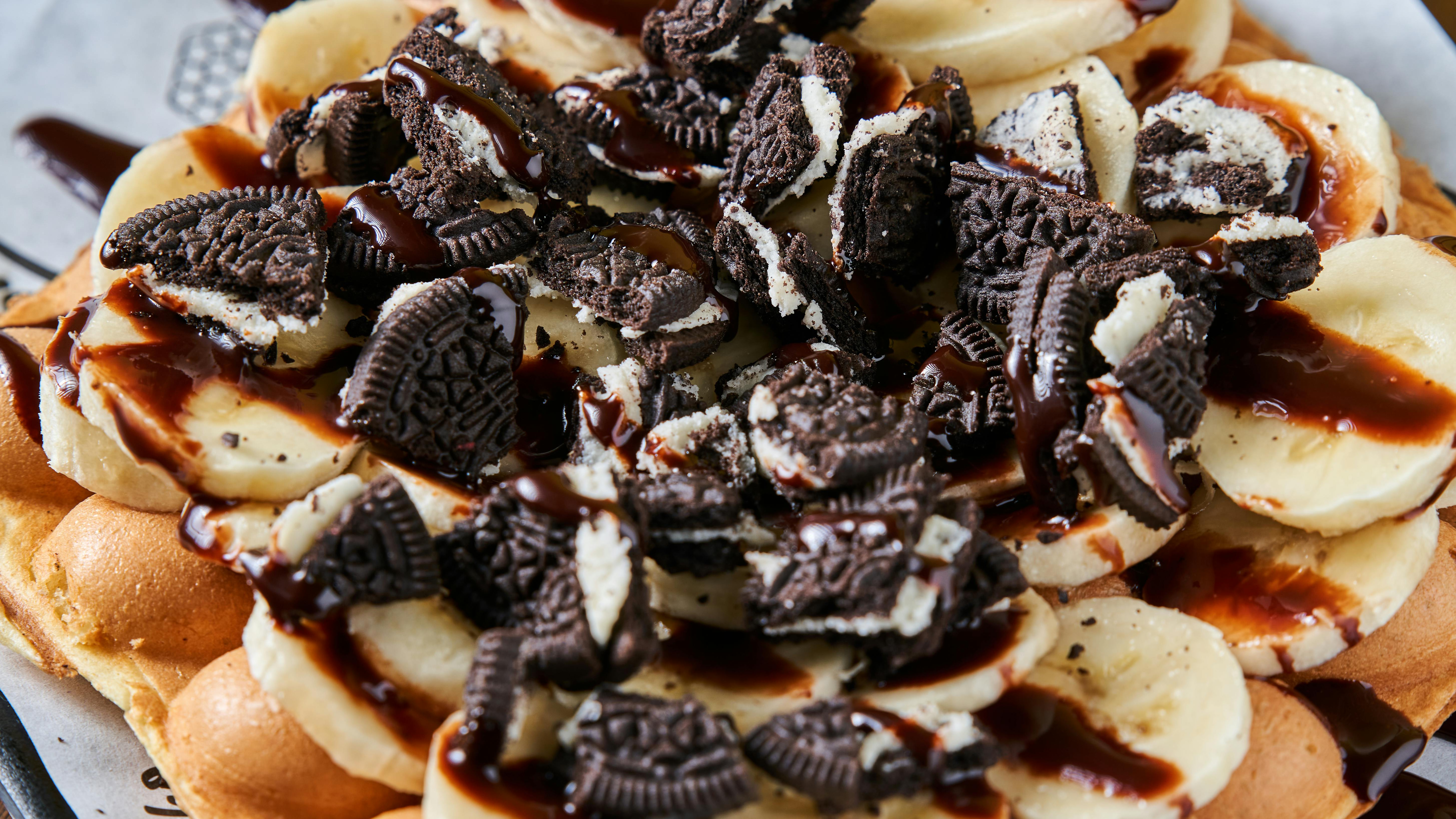 Close-up of a waffle topped with banana slices, crushed cookies, and chocolate sauce.