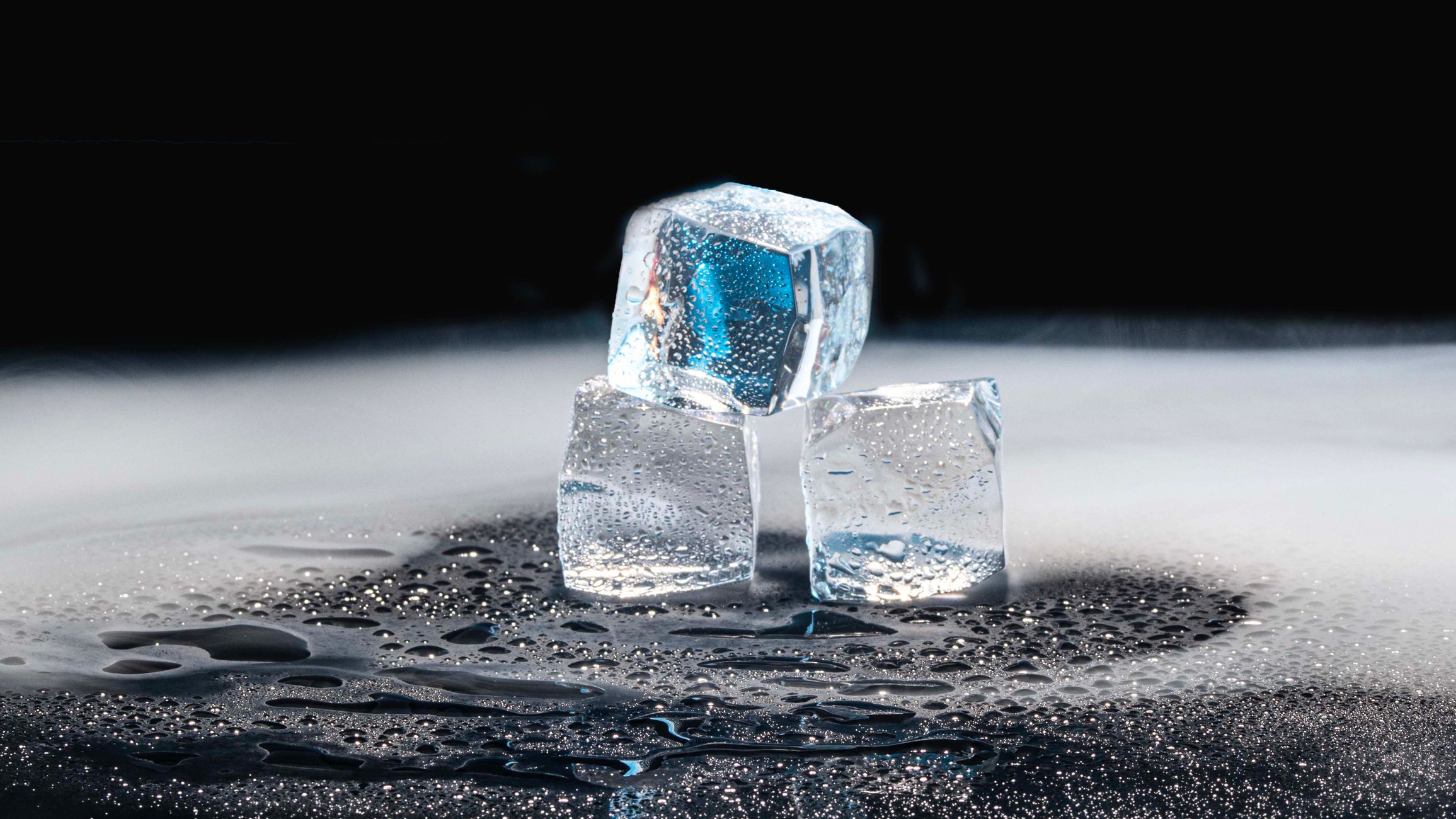 Three ice cubes melting on a black surface with water droplets.