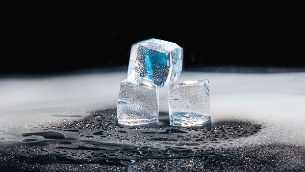Three ice cubes melting on a black surface with water droplets.