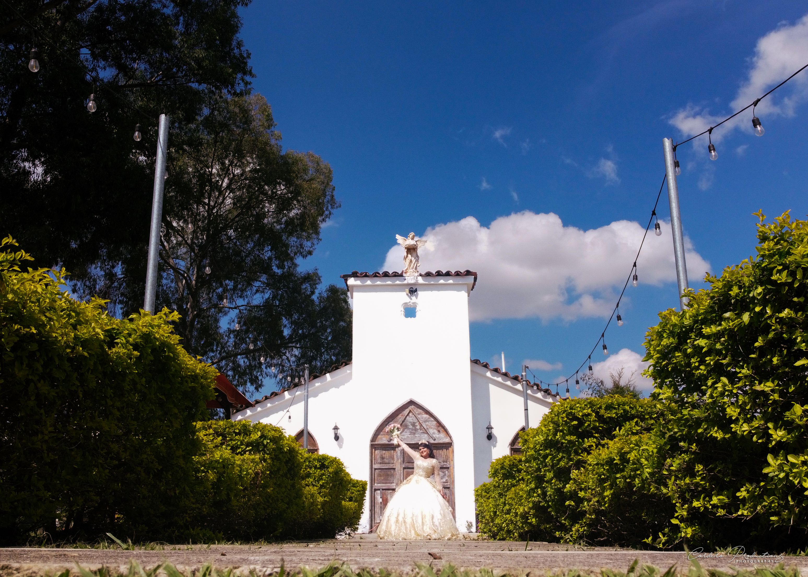 Bride in white dress poses elegantly outside a picturesque chapel under clear blue sky.
