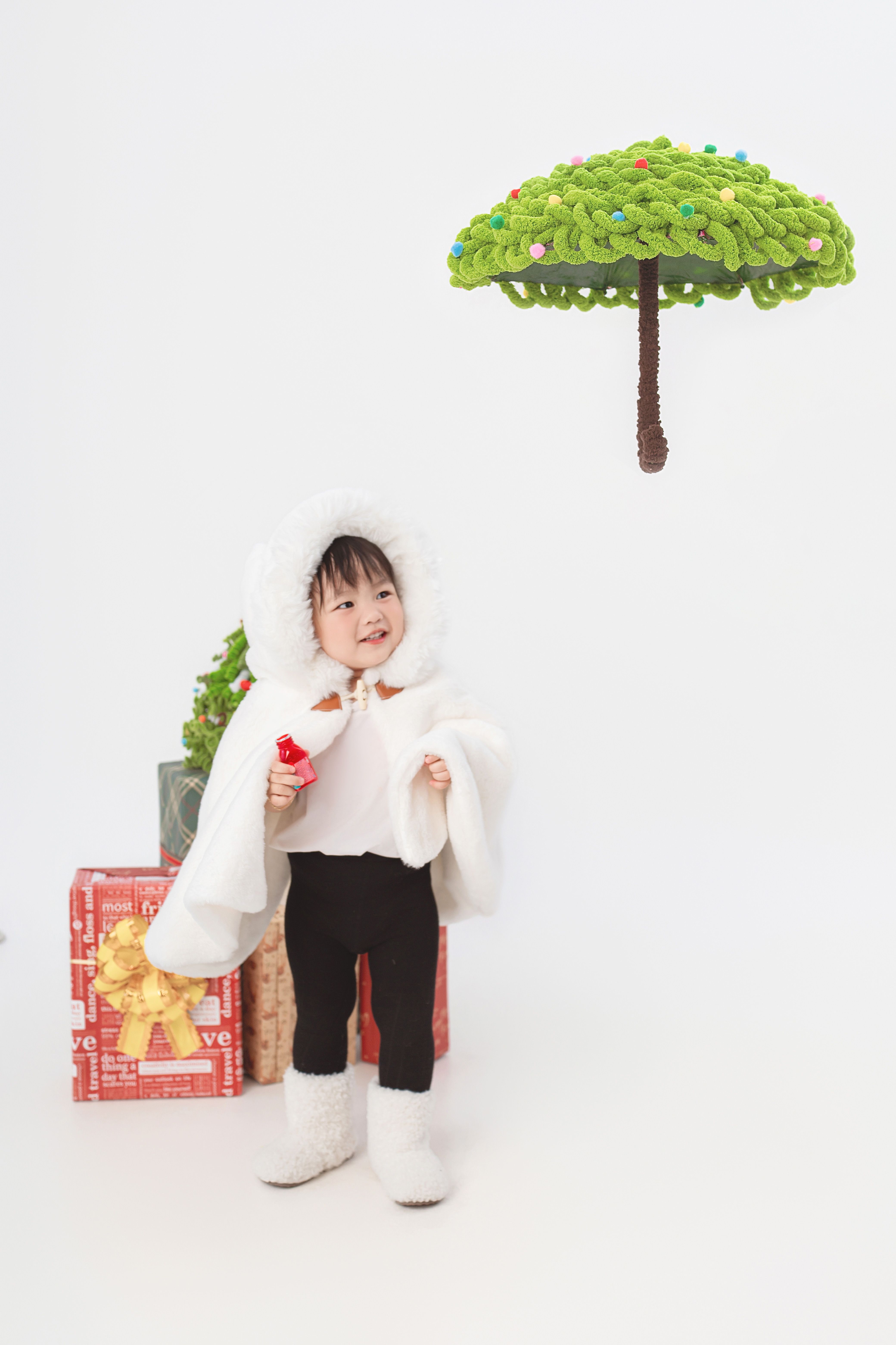 Cute child in hooded outfit standing by gifts with festive decor, smiling joyfully.