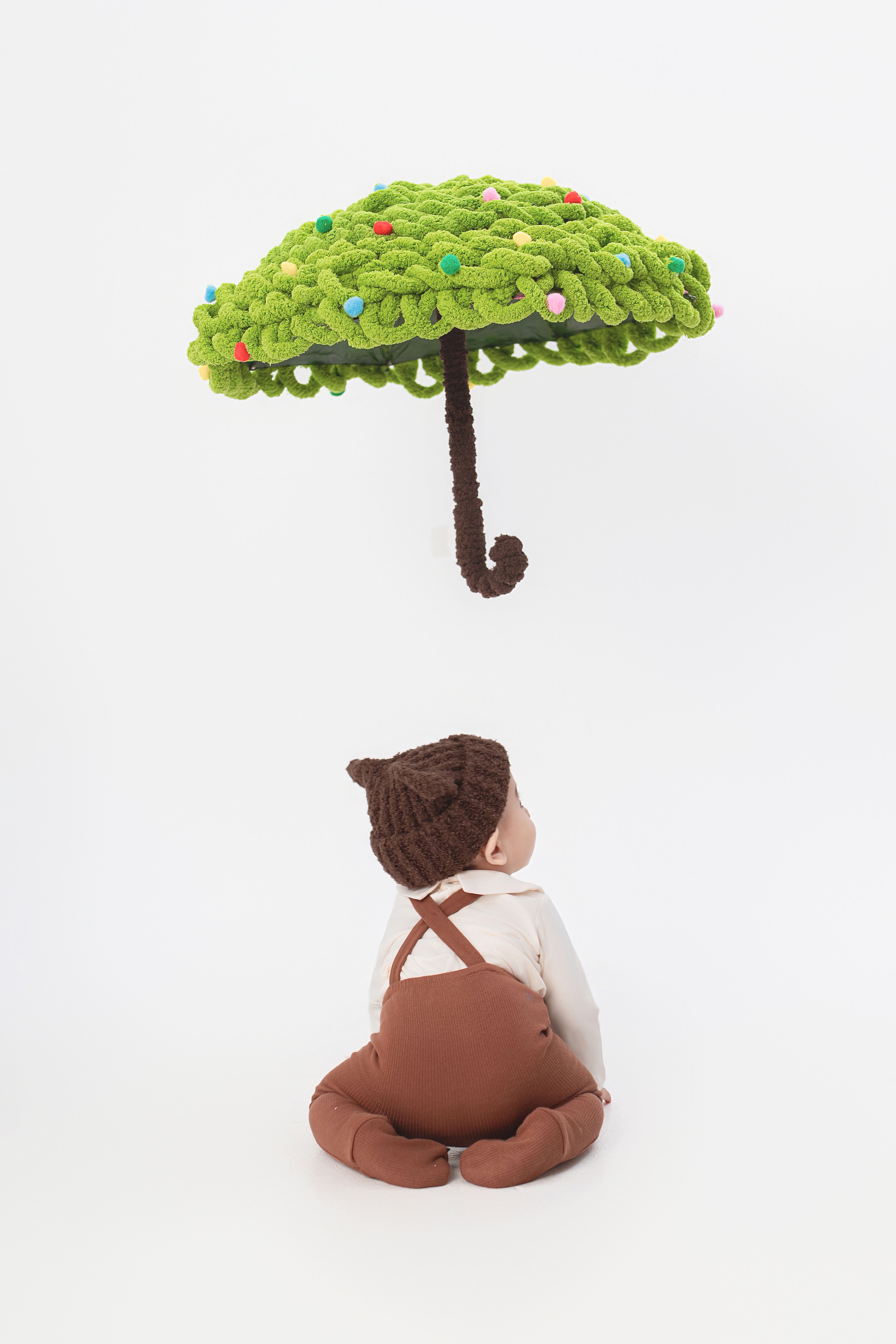 Cute baby in brown outfit gazing up at a fun, knitted green umbrella on a white background.