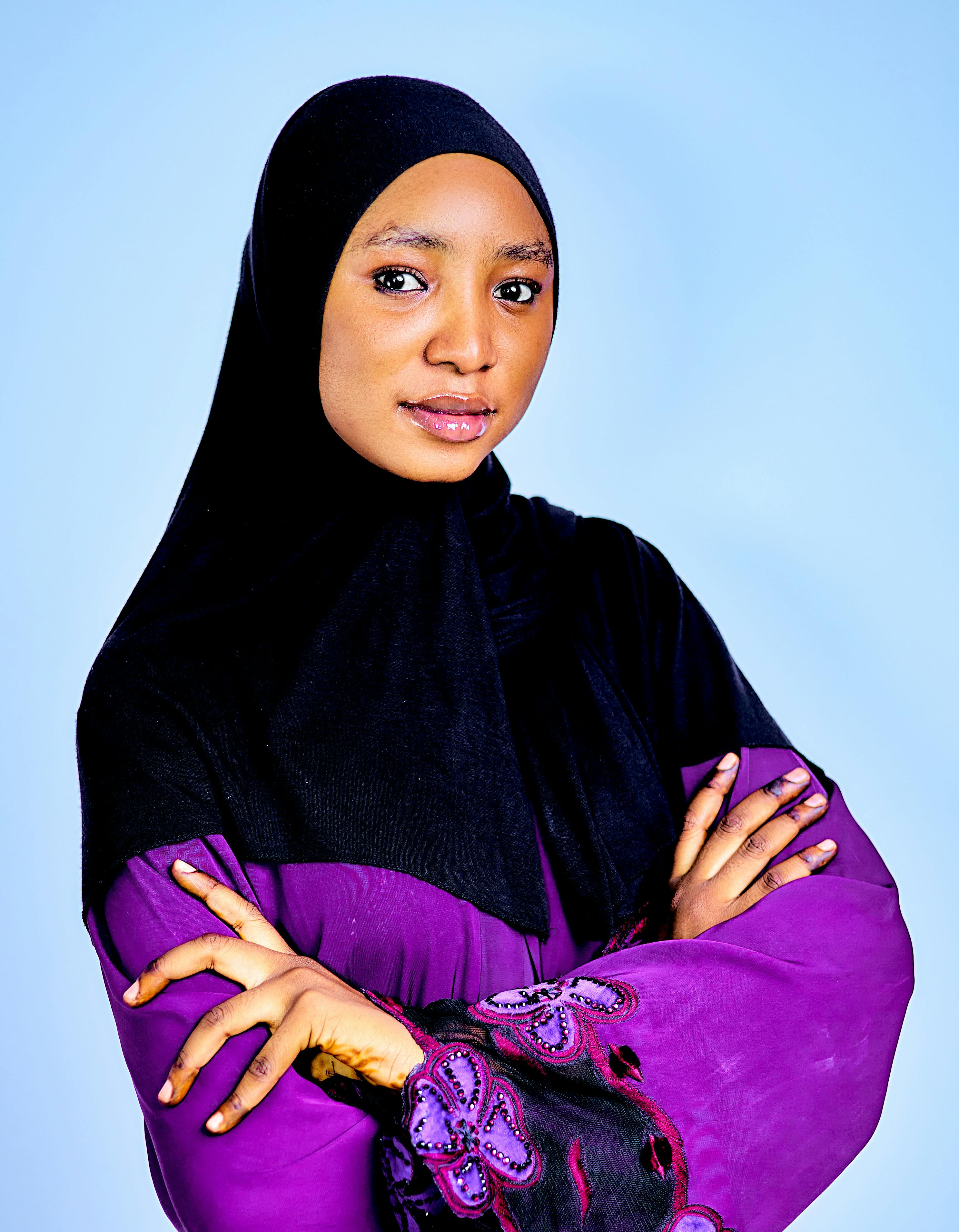 Portrait of a confident woman in a hijab and traditional attire with a light blue background.