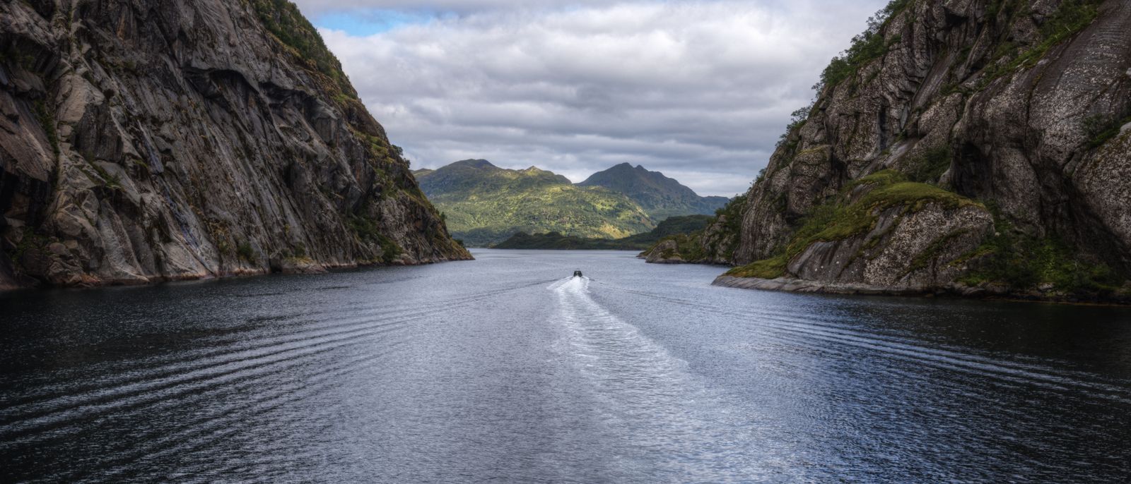 A small vessel moving up a narrow Norwegian fjord between sheer dark rock walls.