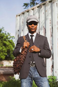 Fashionable man wearing a suit and cap, holding a bag in an outdoor setting.