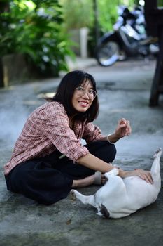 Smiling young woman petting a small dog while sitting on the ground outdoors.