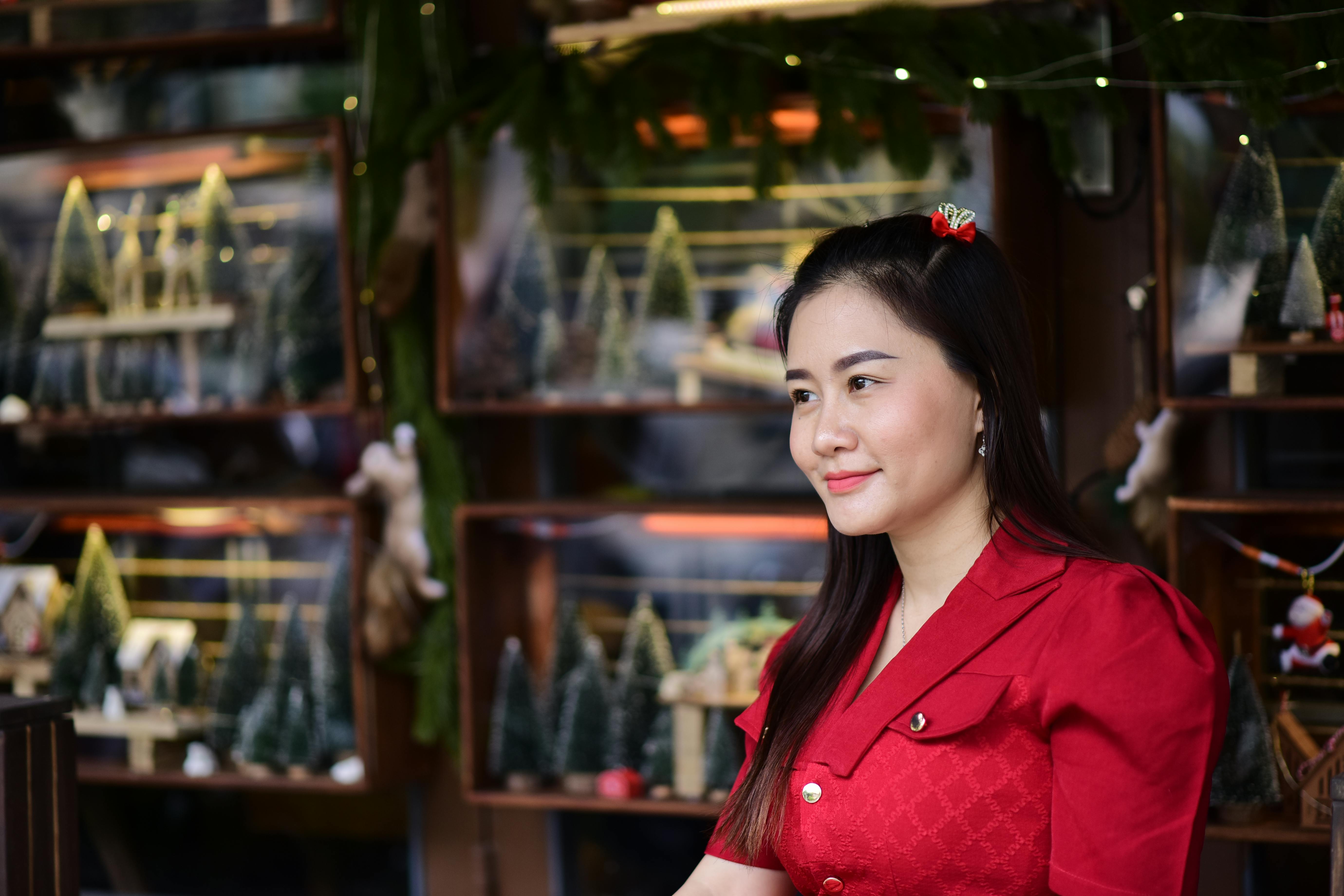 Woman in a bright red dress smiling outdoors by a festive display with small Christmas trees.