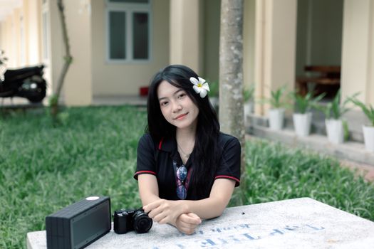 Smiling woman with flower in hair, sitting outdoors with camera.