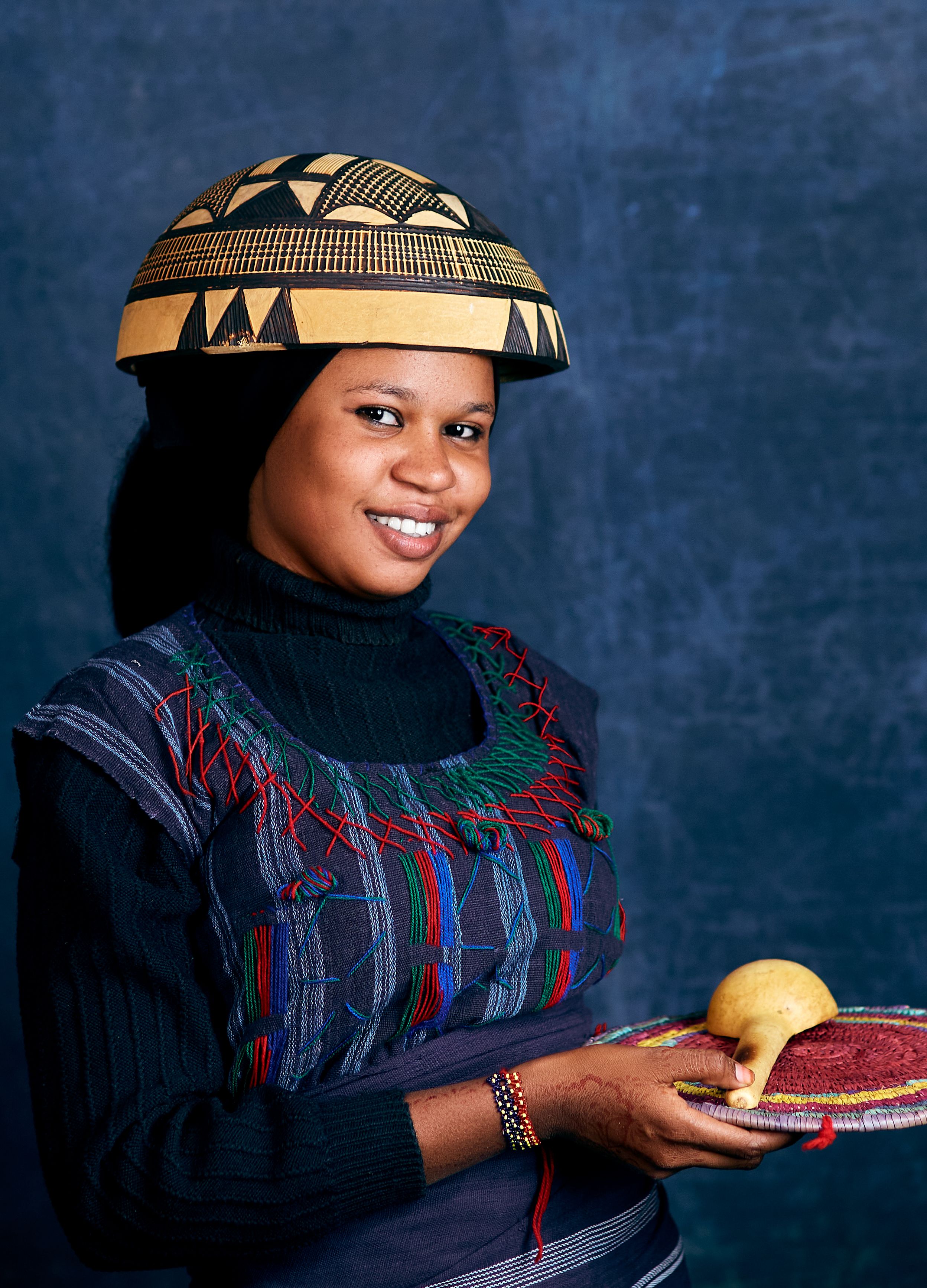 Smiling African woman in traditional attire holding a gourd against a blue background.