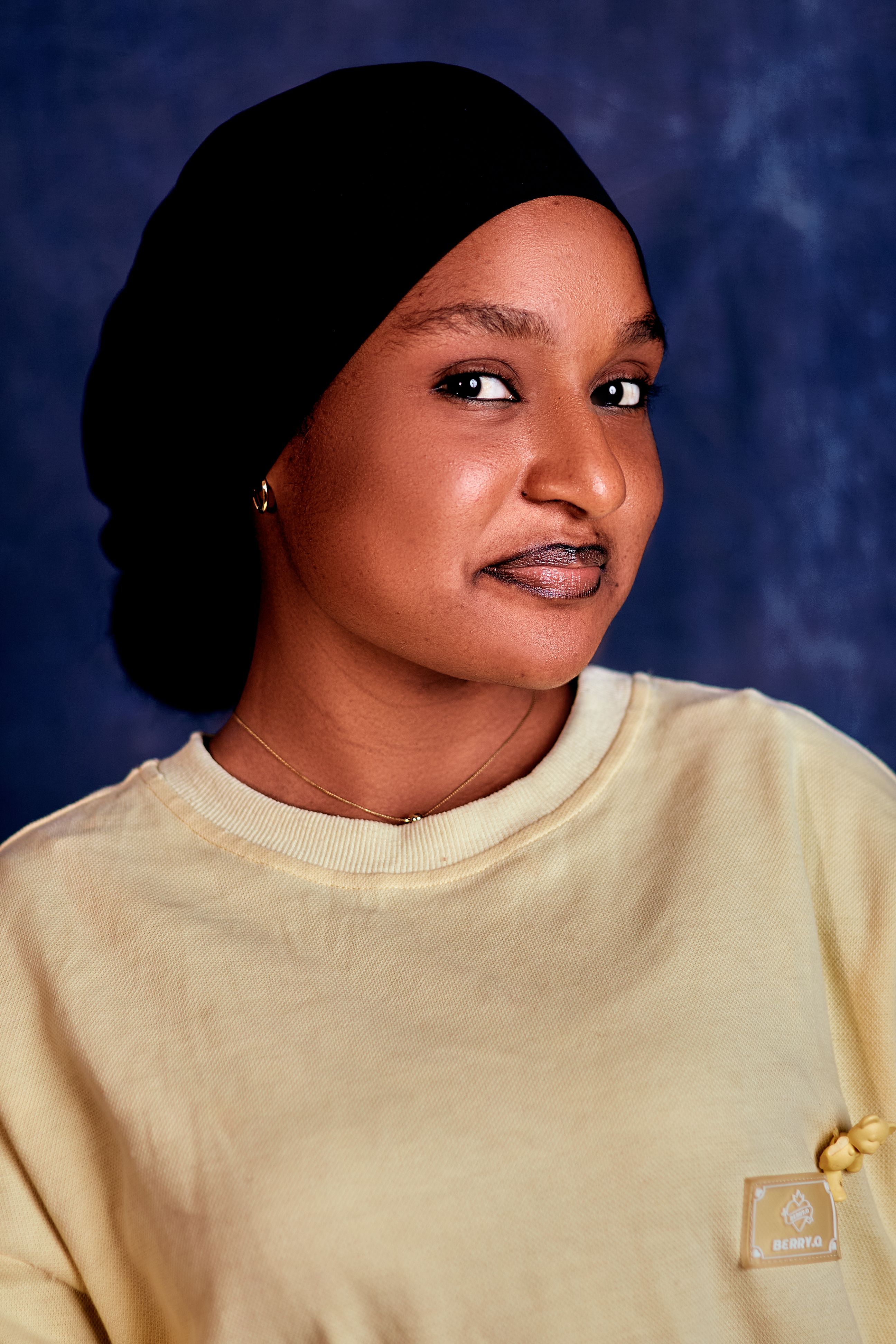 Close-up portrait of a smiling woman wearing a yellow sweater and black headscarf against a dark background.