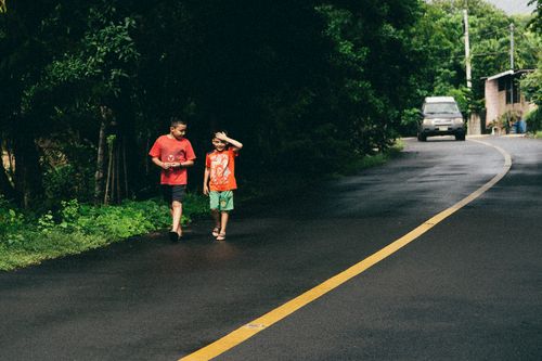 Free Two children walking and chatting on a rural road surrounded by green foliage and a passing car. Stock Photo