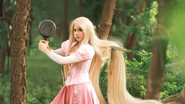 Costumed woman with long blonde hair in a forest setting, holding a pan.