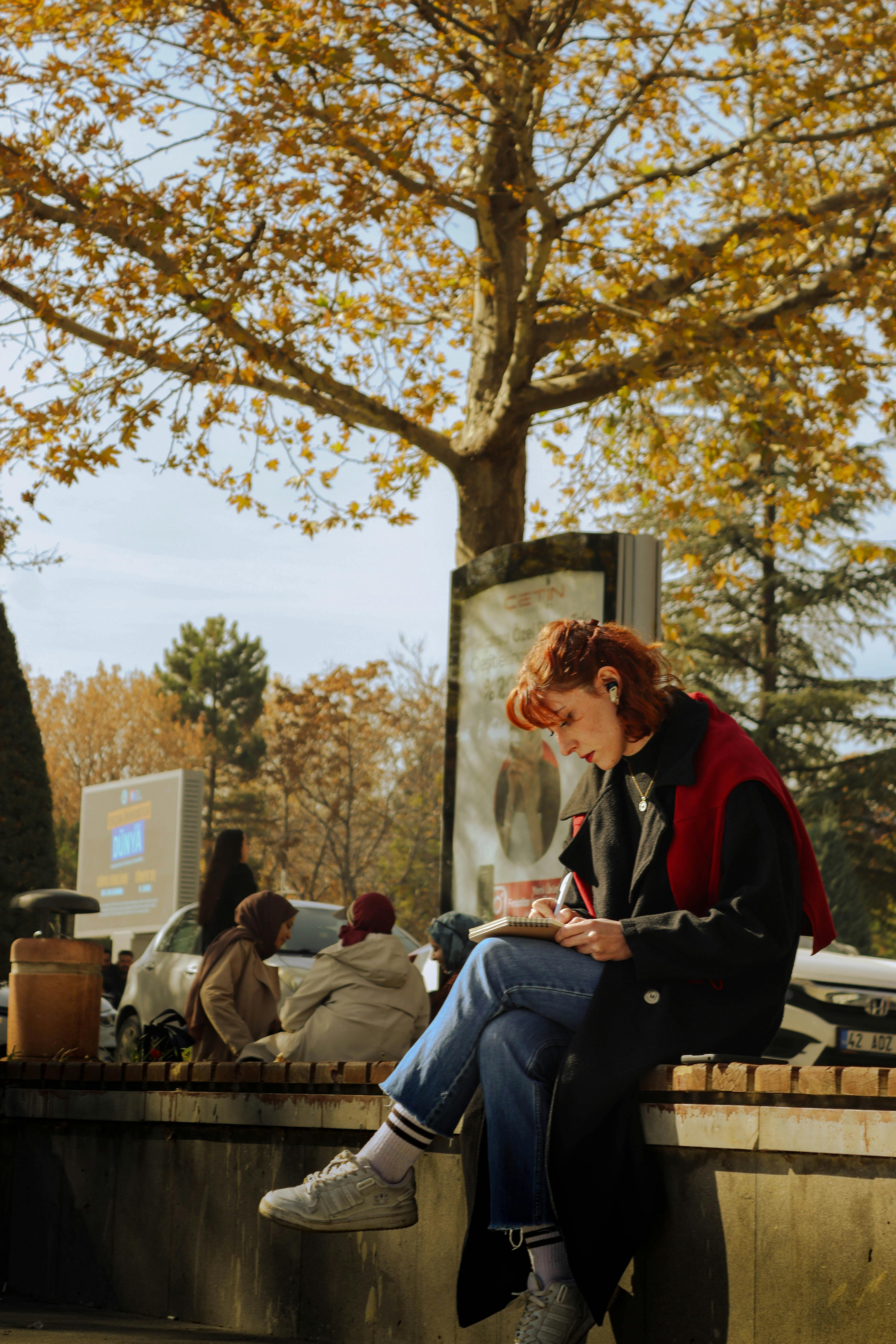 A woman sits reading under golden autumn trees in a serene park setting.