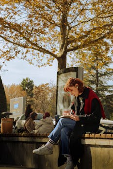 A woman sits reading under golden autumn trees in a serene park setting.