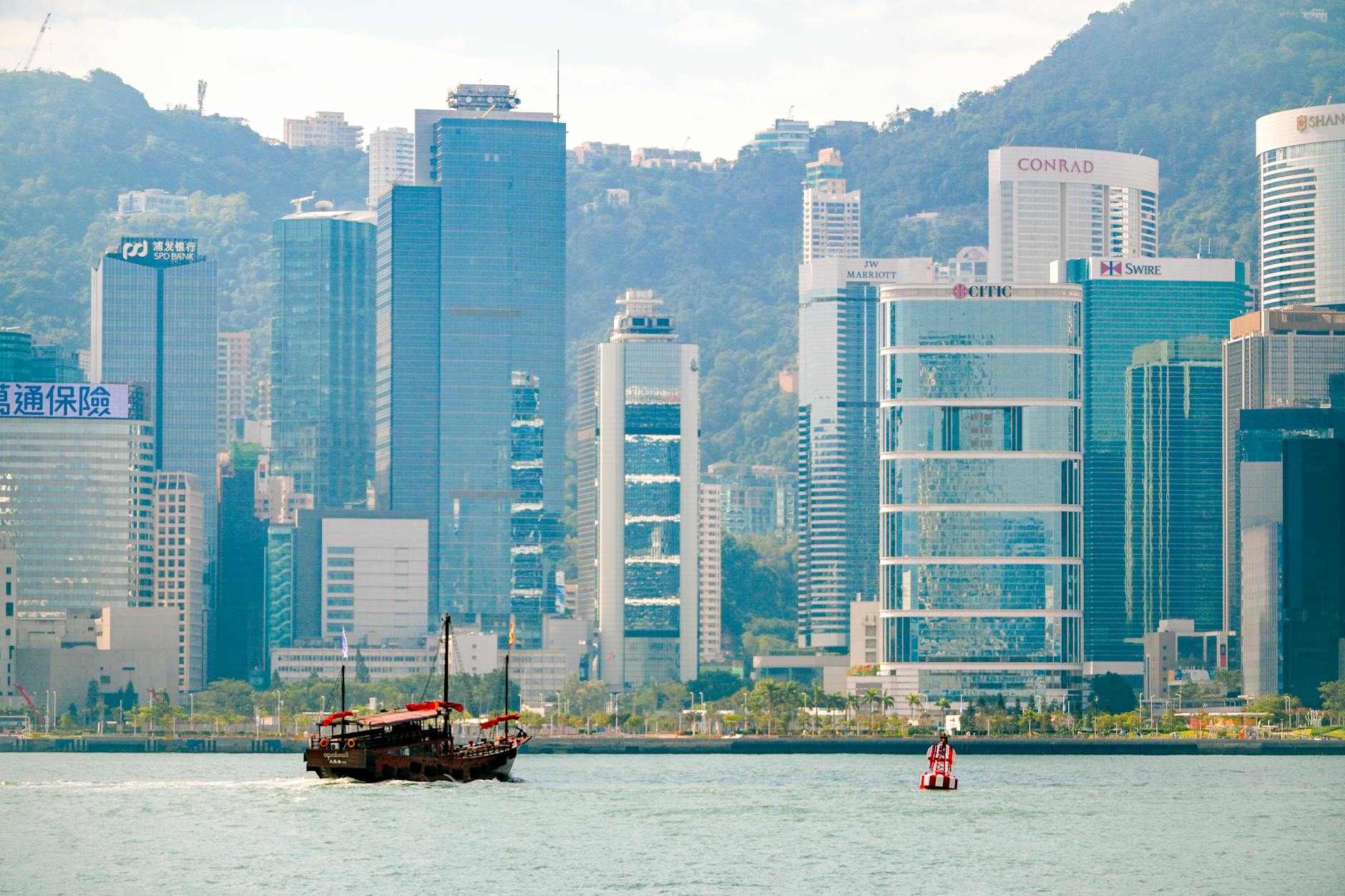 A traditional junk boat sails in front of Hong Kong's iconic skyline, featuring modern skyscrapers.