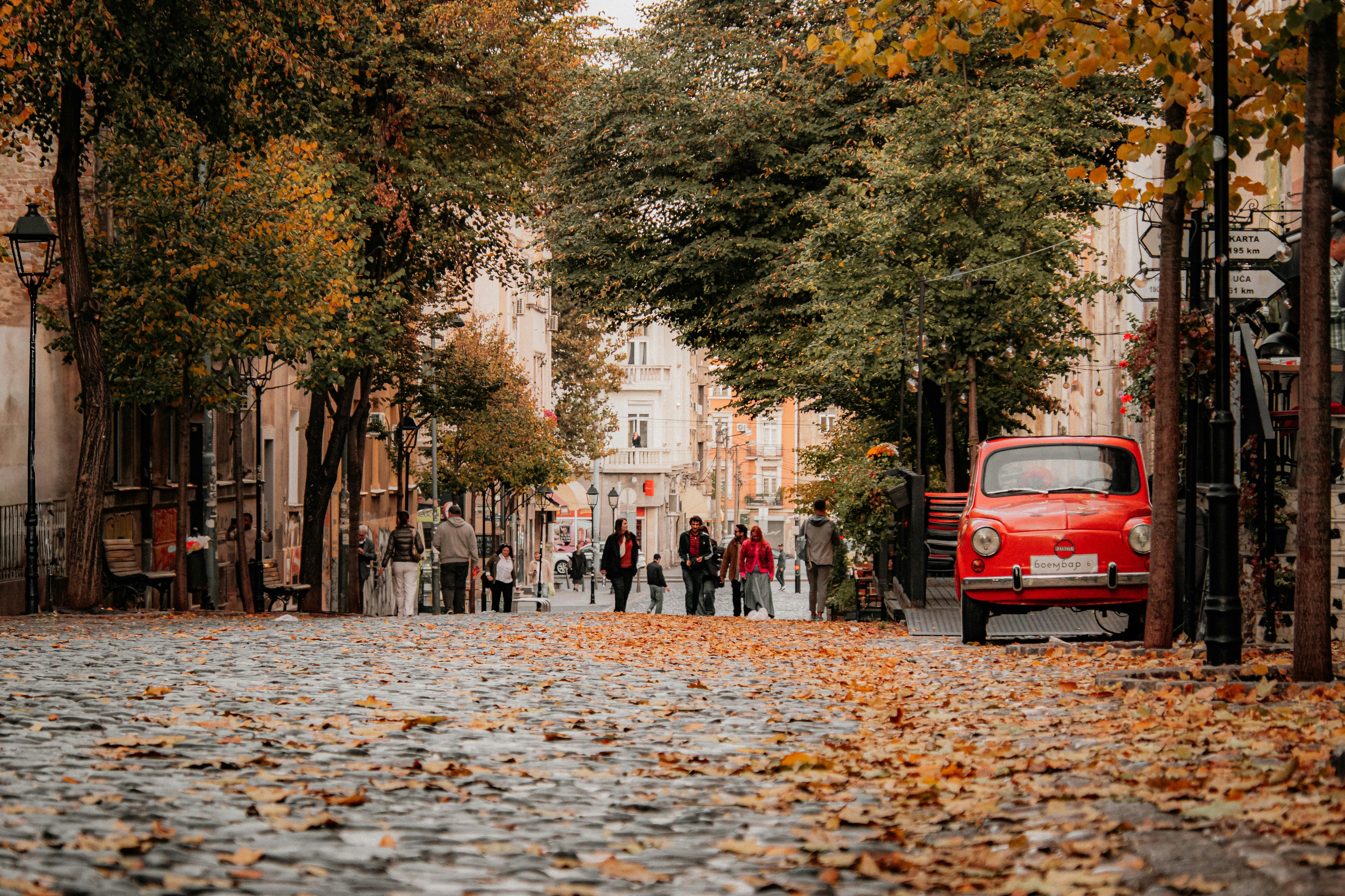 Autumn street scene featuring a vintage red car, fallen leaves, and people strolling along a cobblestone path.