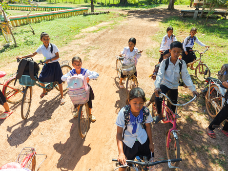 A group of Cambodian children ride bicycles to school on a sunny day, showcasing rural life.