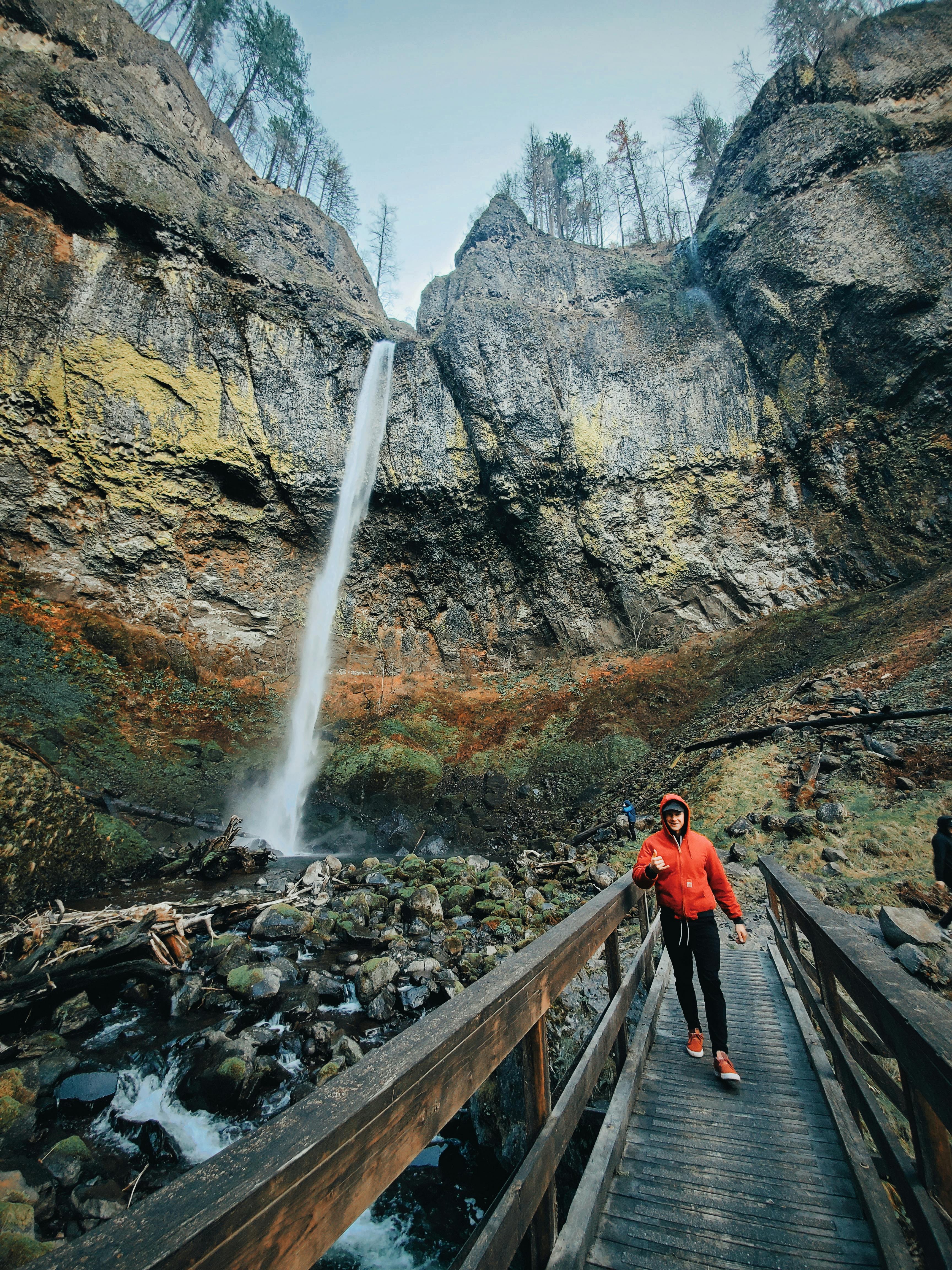 一名徒步旅行者穿着红色夹克，穿过人行天桥，背景是一座壮观的瀑布