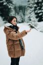 一位穿着棕色大衣、面带微笑的女子在白雪皑皑的森林中抱着一只小鸟，展现出宁静的冬日户外场景