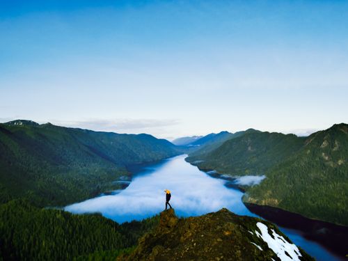 Free A breathtaking view of a lone hiker standing at a mountain peak overlooking a vast, scenic valley and river. Stock Photo