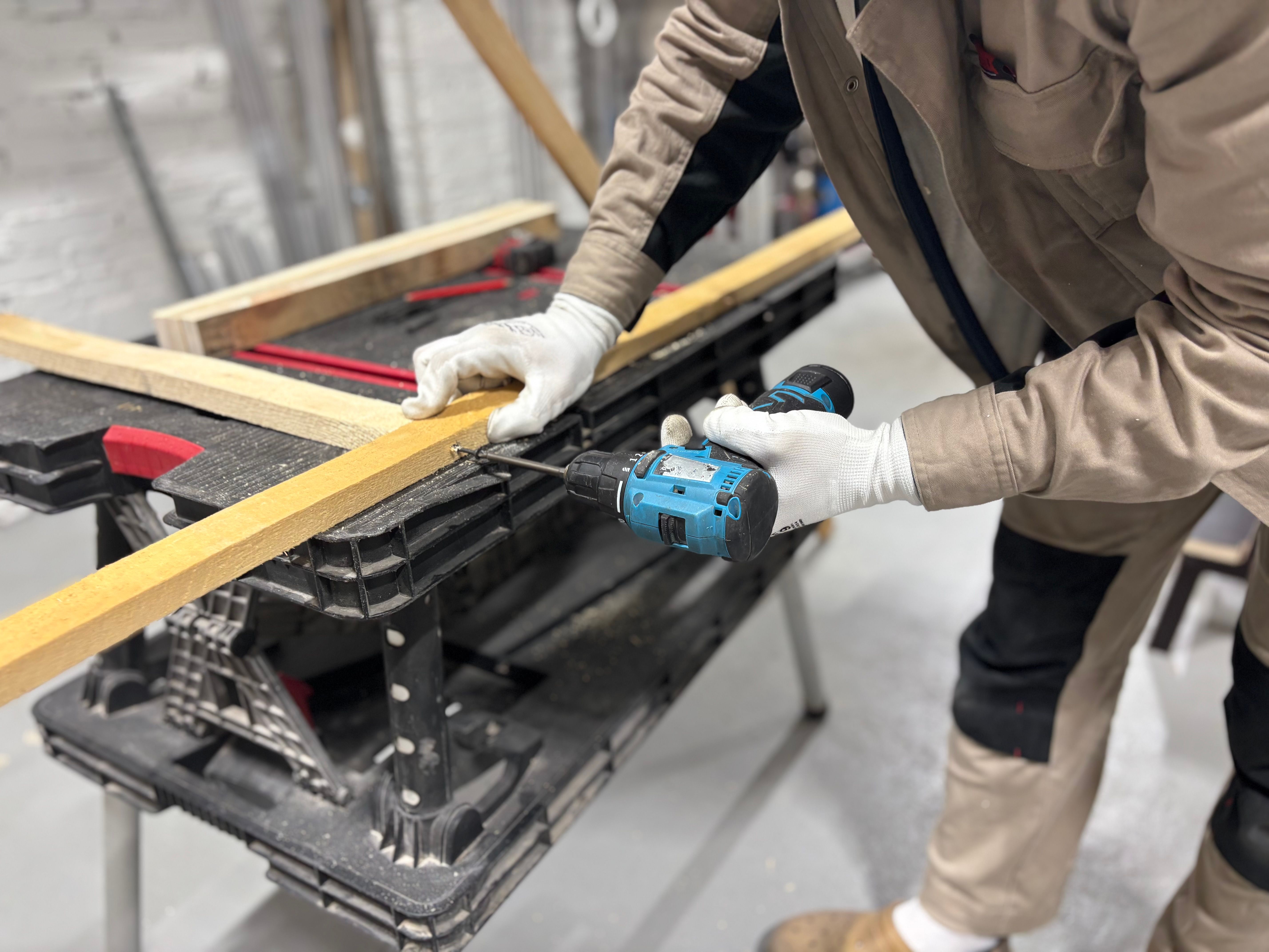 Carpenter wearing gloves using a cordless drill on a wooden plank at a workshop bench