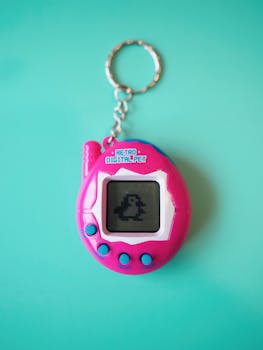 Bright pink retro digital pet keychain with pixel art display on a turquoise background.