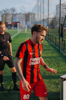 A football player in a red and black jersey on the field during a sunny day match.