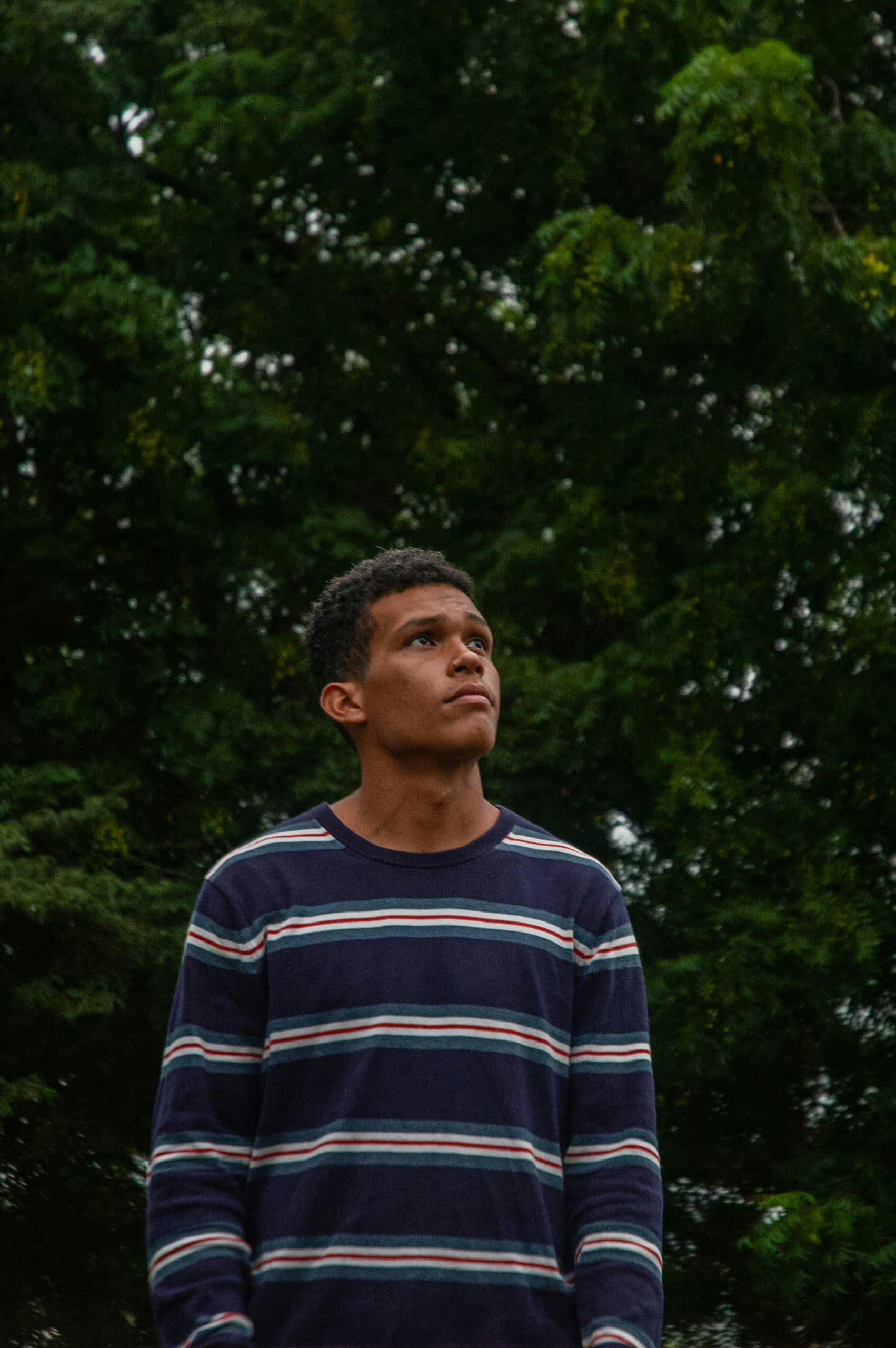 Gratis Un giovane che indossa un maglione a righe è in piedi, pensieroso, davanti a rigogliosi alberi verdi. Foto a disposizione