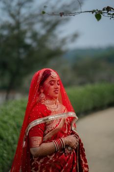 Traditional Indian bride in red saree and jewelry outdoors, serene expression.