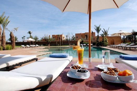 Enjoy a luxurious poolside scene with sun loungers, palm trees, and refreshing drinks under a bright sky.