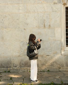A woman in a leather jacket takes photos against a stone wall in a daylight setting.