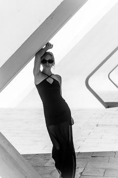 Elegant woman in black dress leaning against modern architecture structure.