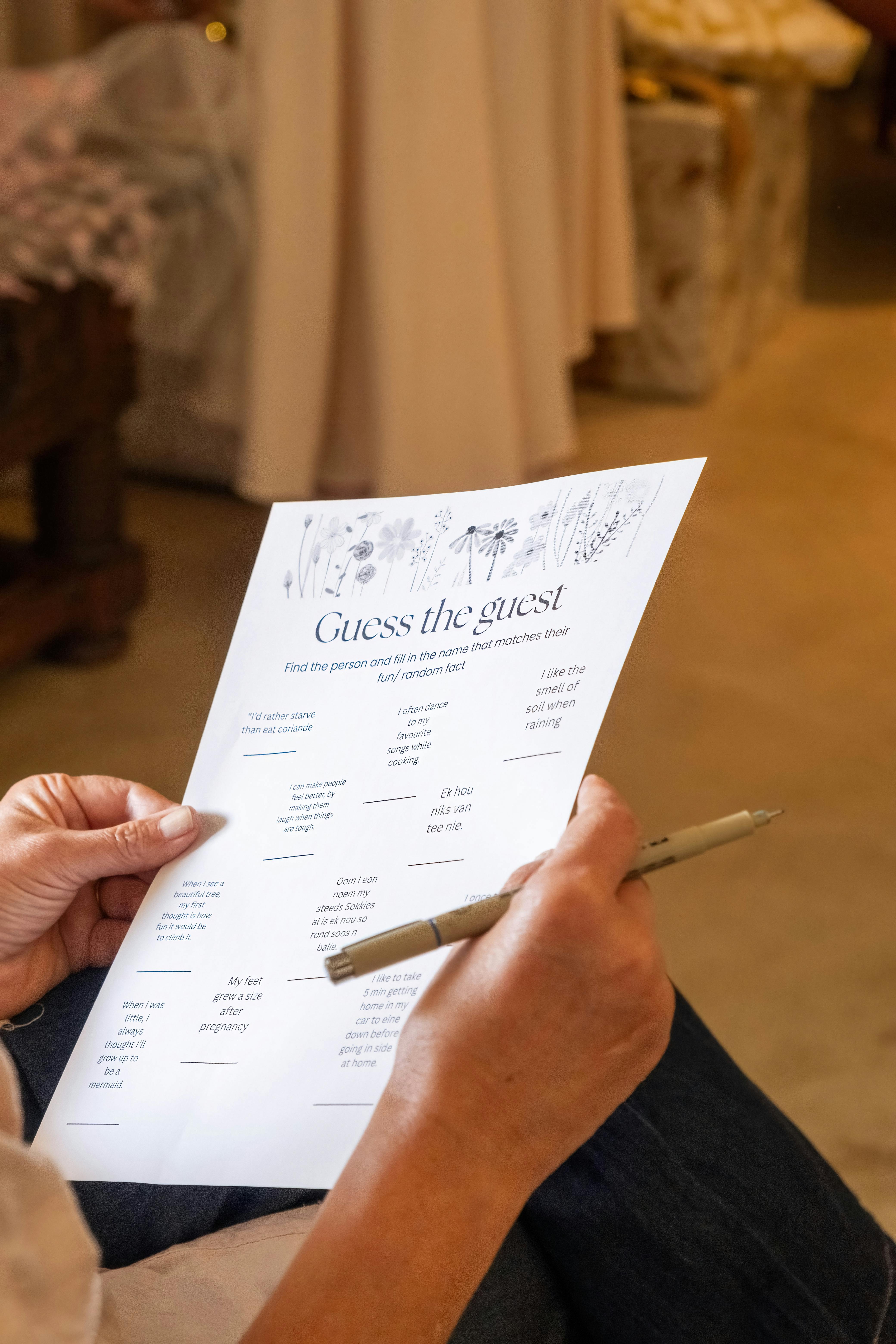 A person holds a pen and a 'Guess the Guest' game paper, engaging in a group activity indoors.