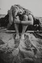 Black and white melancholic young man in casual clothes and headband sitting on rock embracing knees pensively on coastline