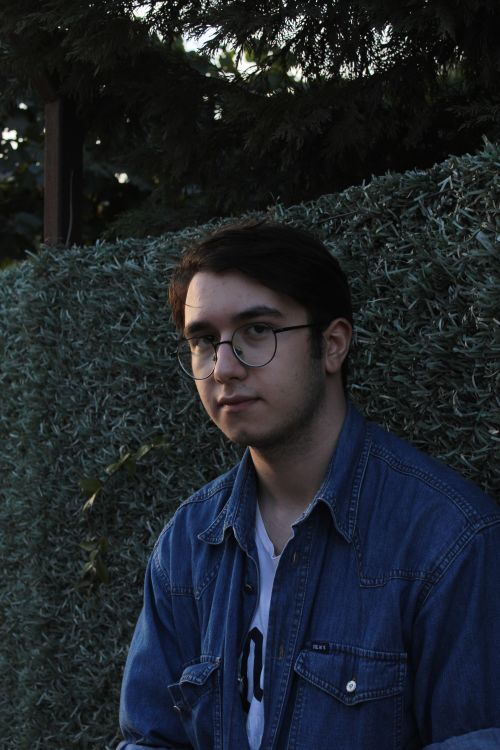 Free Candid portrait of a young man wearing glasses and a denim jacket against a leafy backdrop. Stock Photo