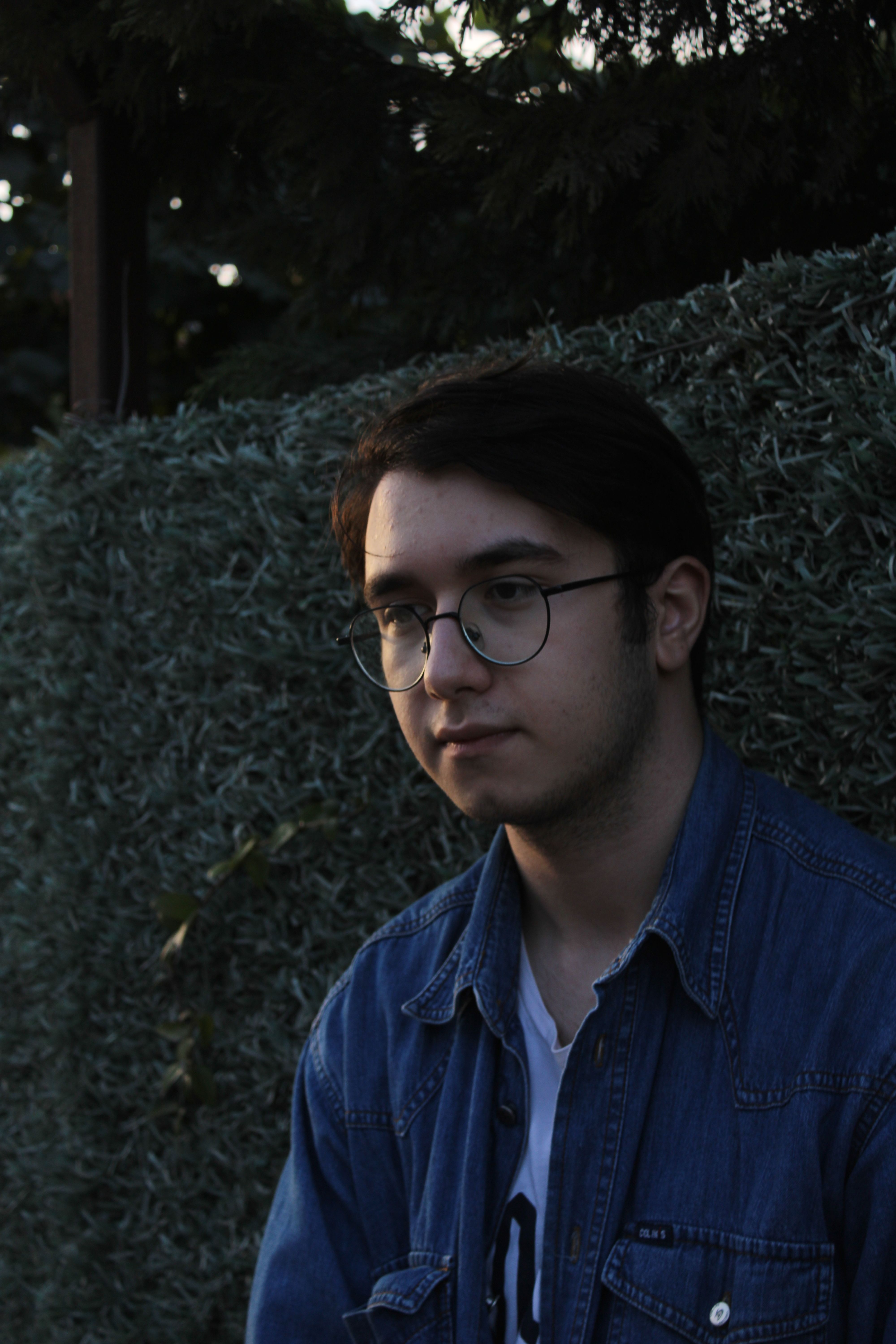 A young man in glasses and denim sits pensively outdoors against a green hedge.