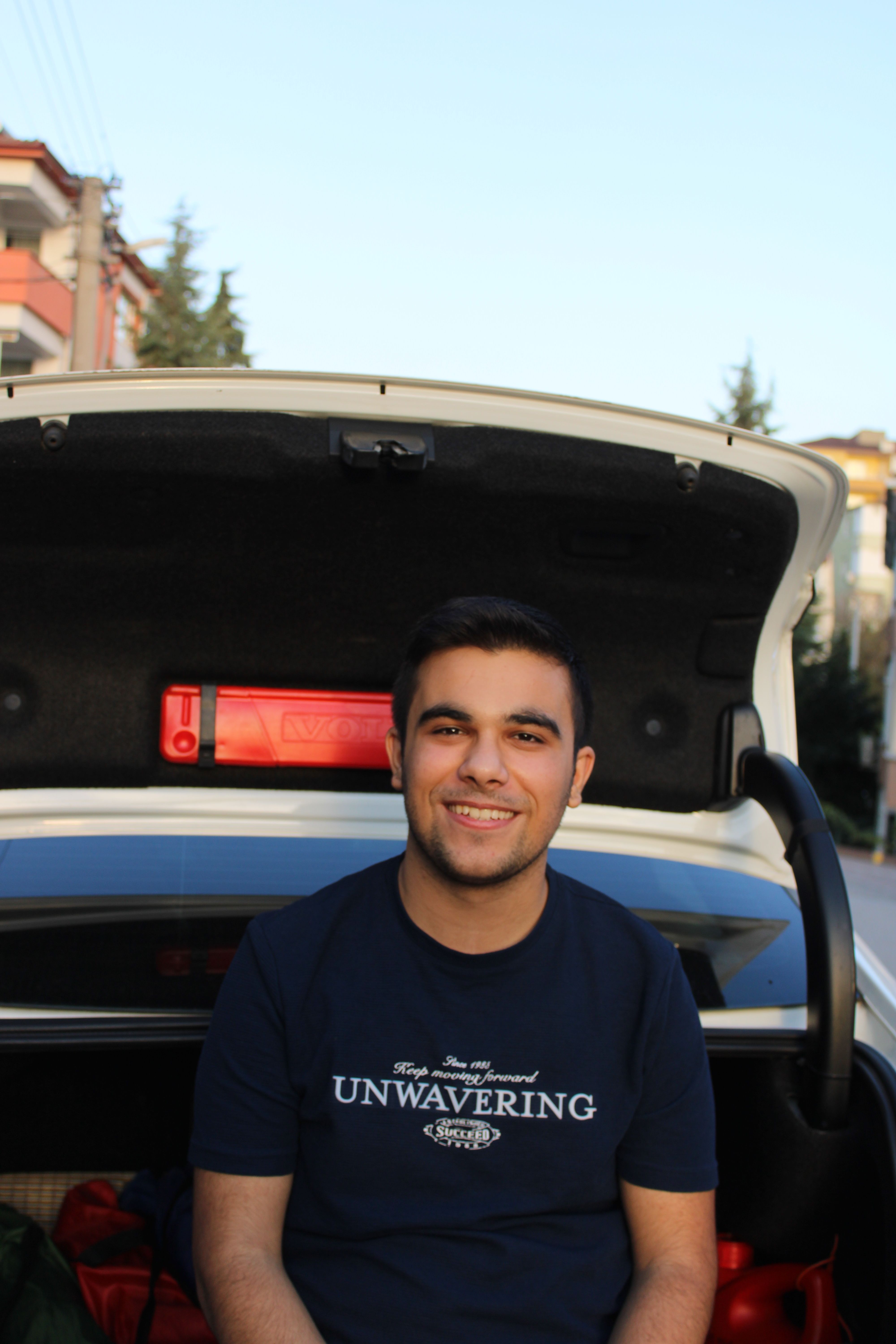 A young man smiling while sitting in an open car trunk on a sunny day in Darıca, Türkiye.
