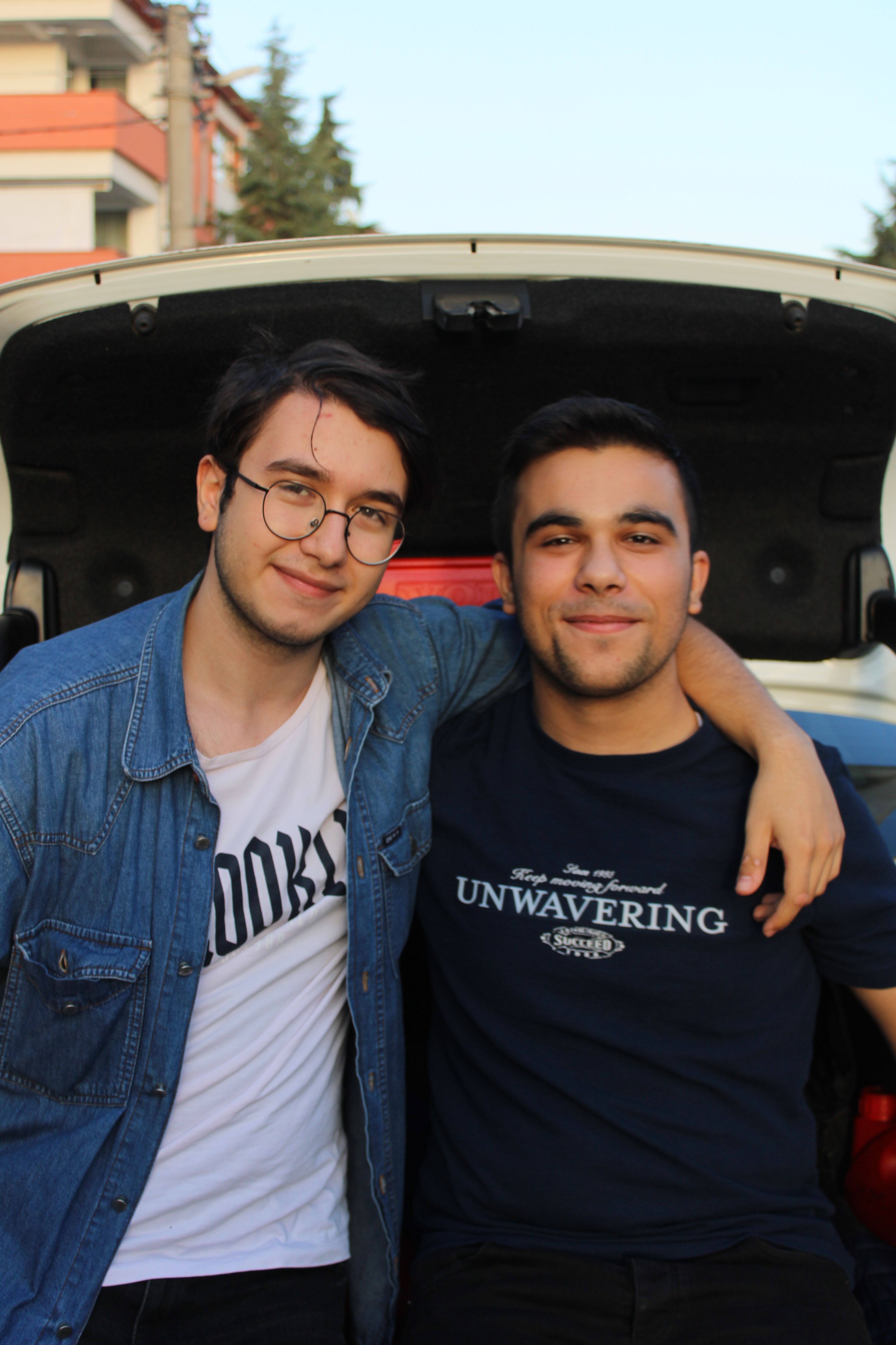 Two young adults smiling and embracing in front of an open car trunk outdoors.