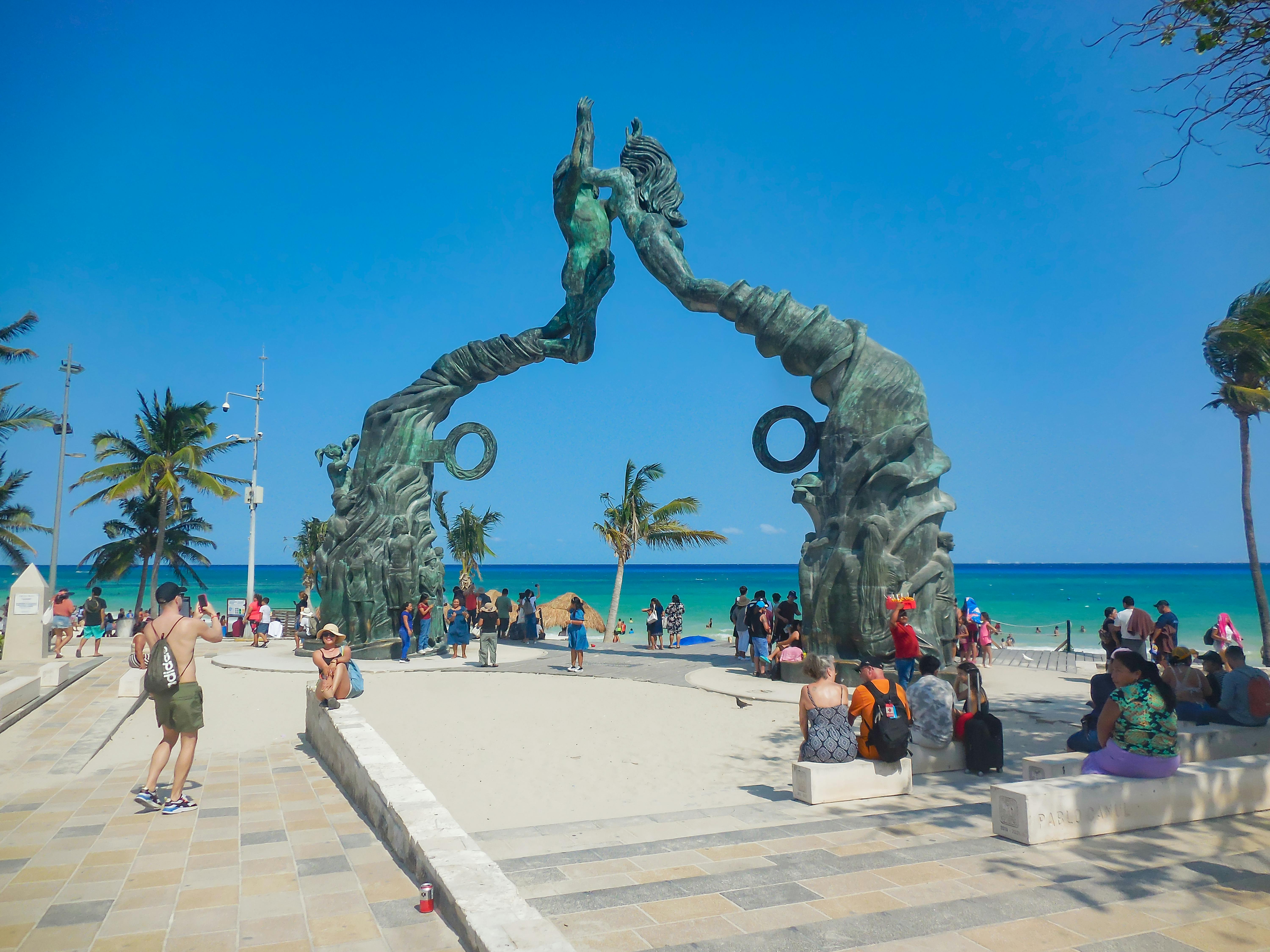 A vibrant scene of tourists at the iconic Portal Maya sculpture in Playa del Carmen, Mexico.