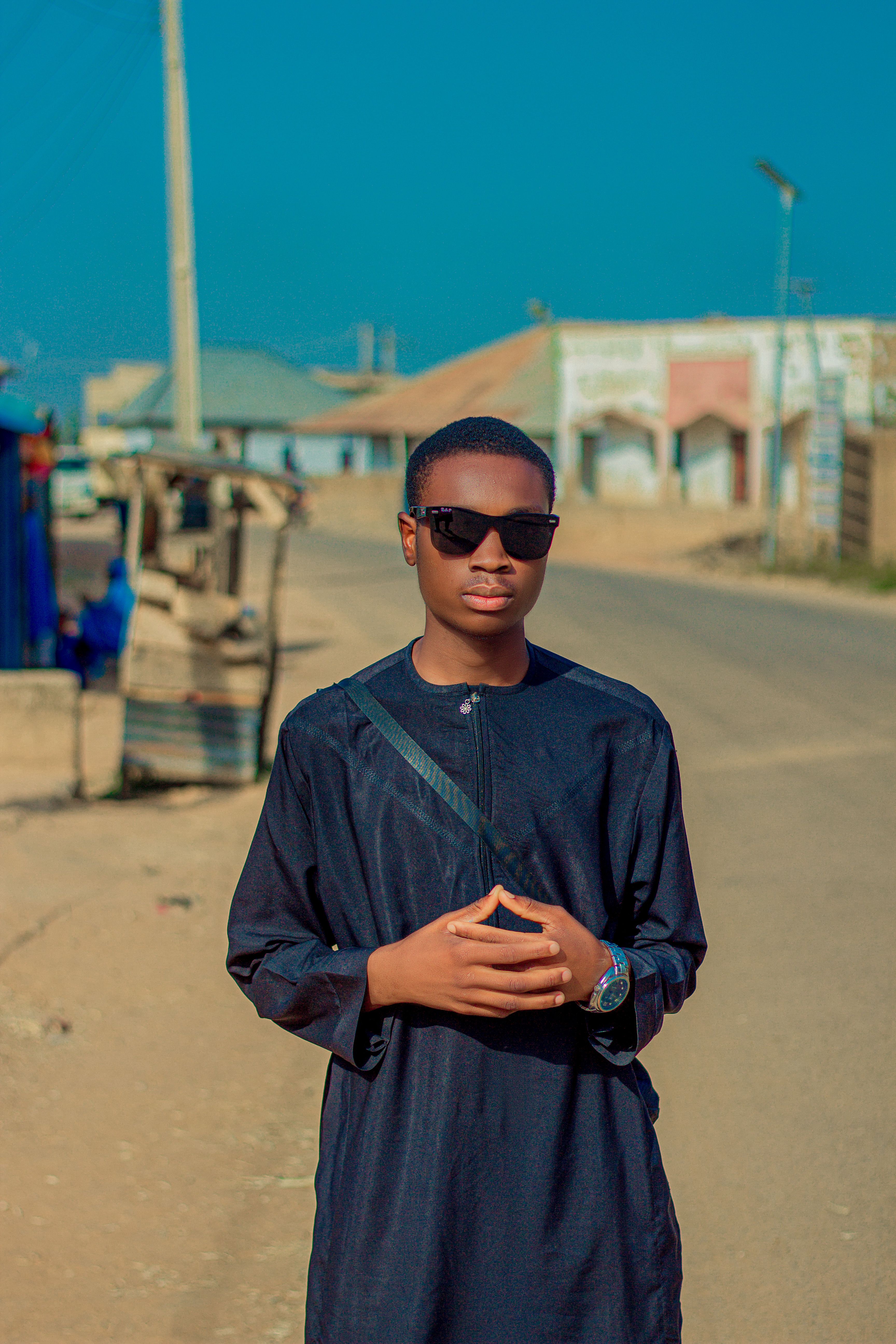 Young man wearing traditional attire with sunglasses standing on a sunny street.
