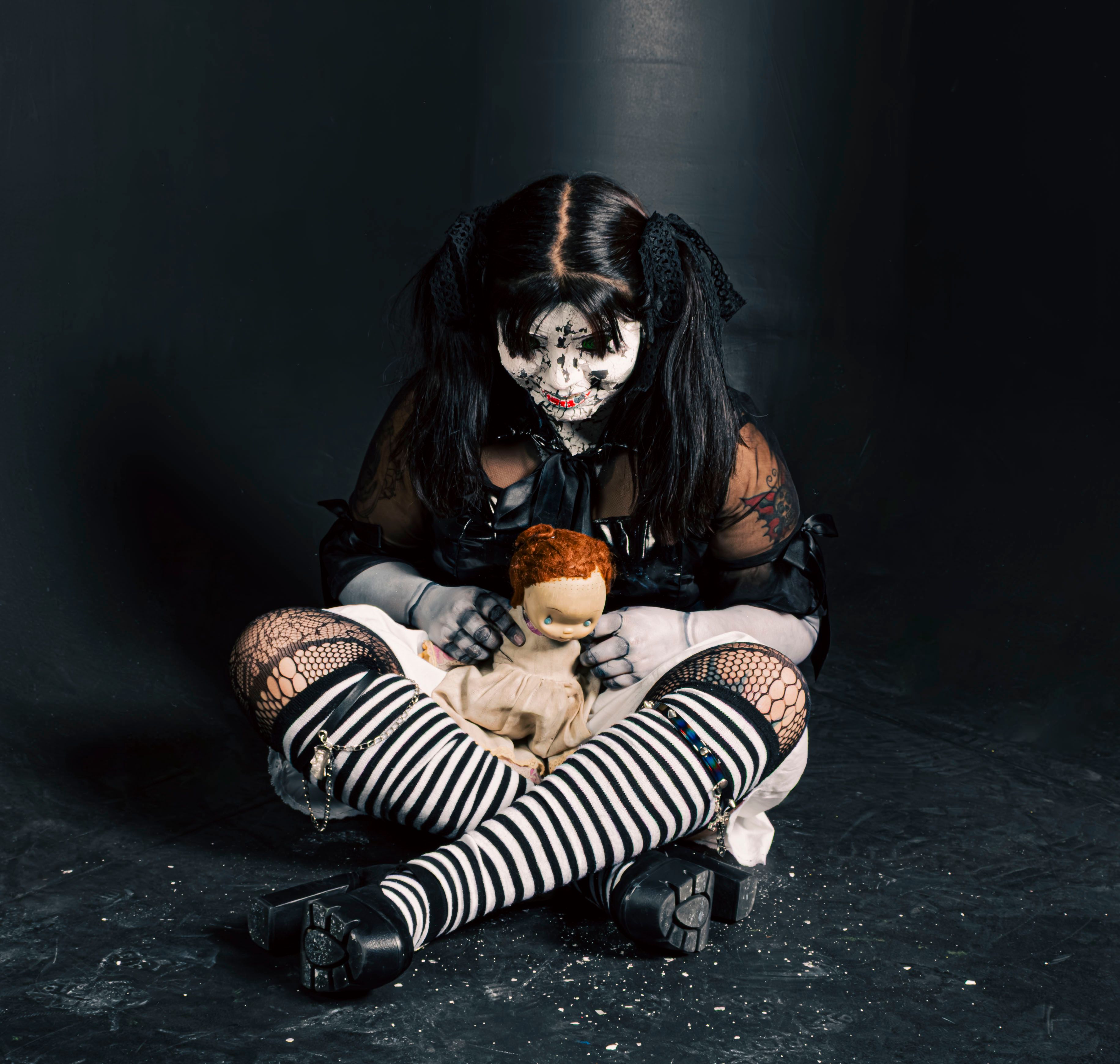 Gothic girl in eerie makeup holding a doll in a dark room.