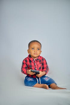 Cute baby boy in a red plaid shirt holding a remote control, sitting on a white background.