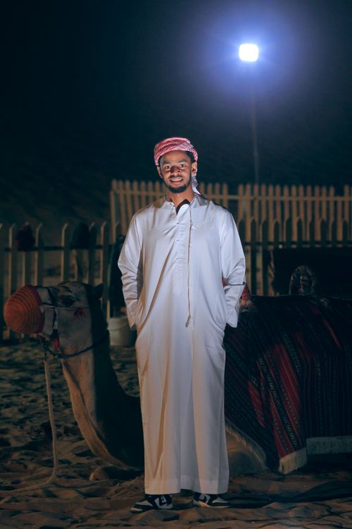 Free A man in traditional attire stands beside a camel during a night desert setting. Stock Photo