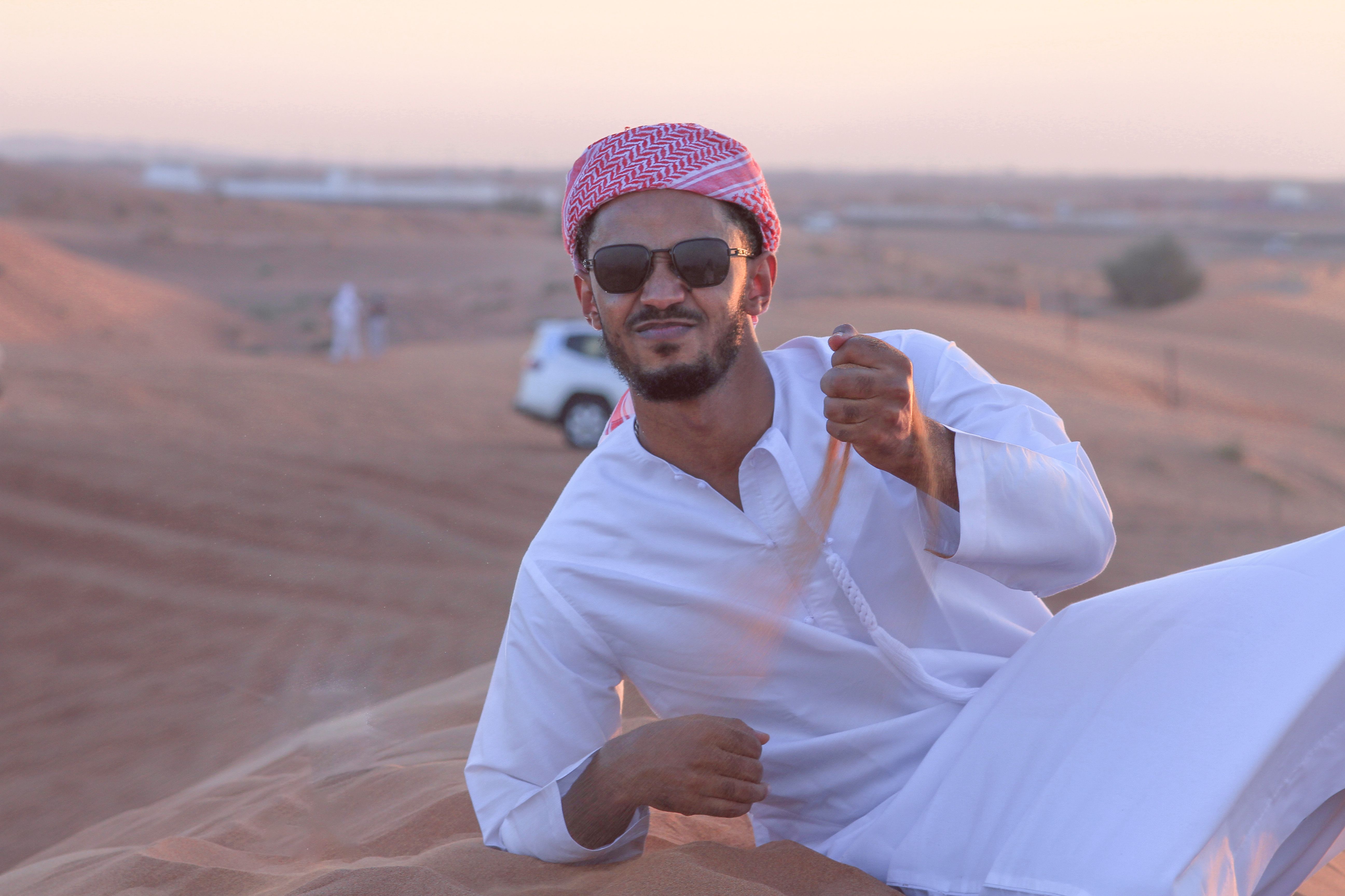 Man wearing traditional Middle Eastern attire relaxing in a desert landscape with a car in the background.